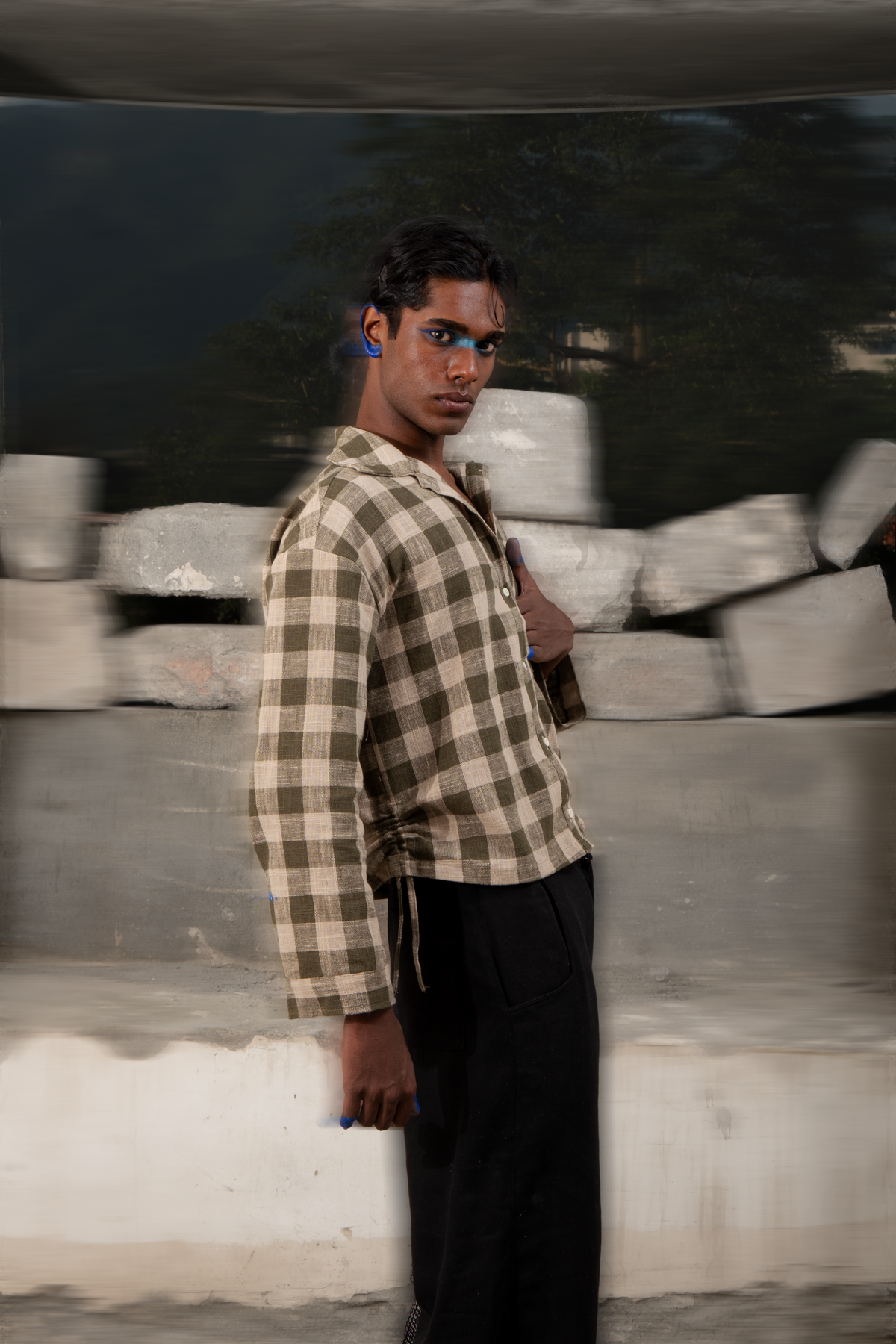 A man wearing a beige and brown checkered long-sleeve shirt and black pants, standing in front of a concrete wall with a reflective dark surface behind him.
